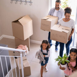 Ethnic family with two children carrying boxes and plant in new home on moving day. High angle view of happy smiling daughters helping mother and father with cardboard boxes in new house. Top view of excited kids having fun walking up stairs running to their rooms while parents holding boxes.